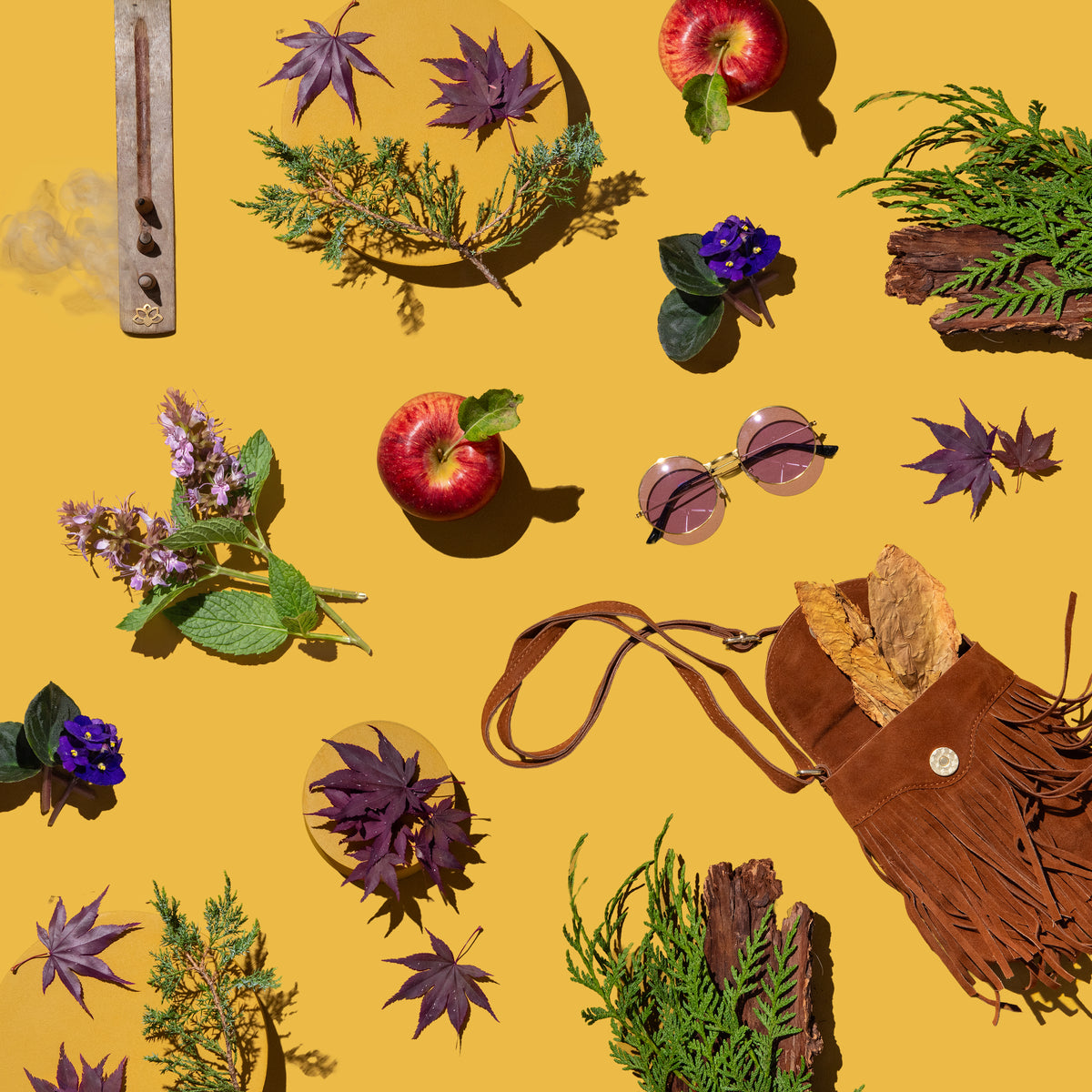 Flat-lay on a mustard-yellow background with scattered botanical and vintage props: purple Japanese maple leaves, juniper and cedar sprigs, red apples, African violets, lavender-pink flowering mint, chunks of tree bark, dried tobacco leaves spilling from a brown suede fringe bag, round rose-tinted sunglasses, and a wooden incense holder with curling smoke.