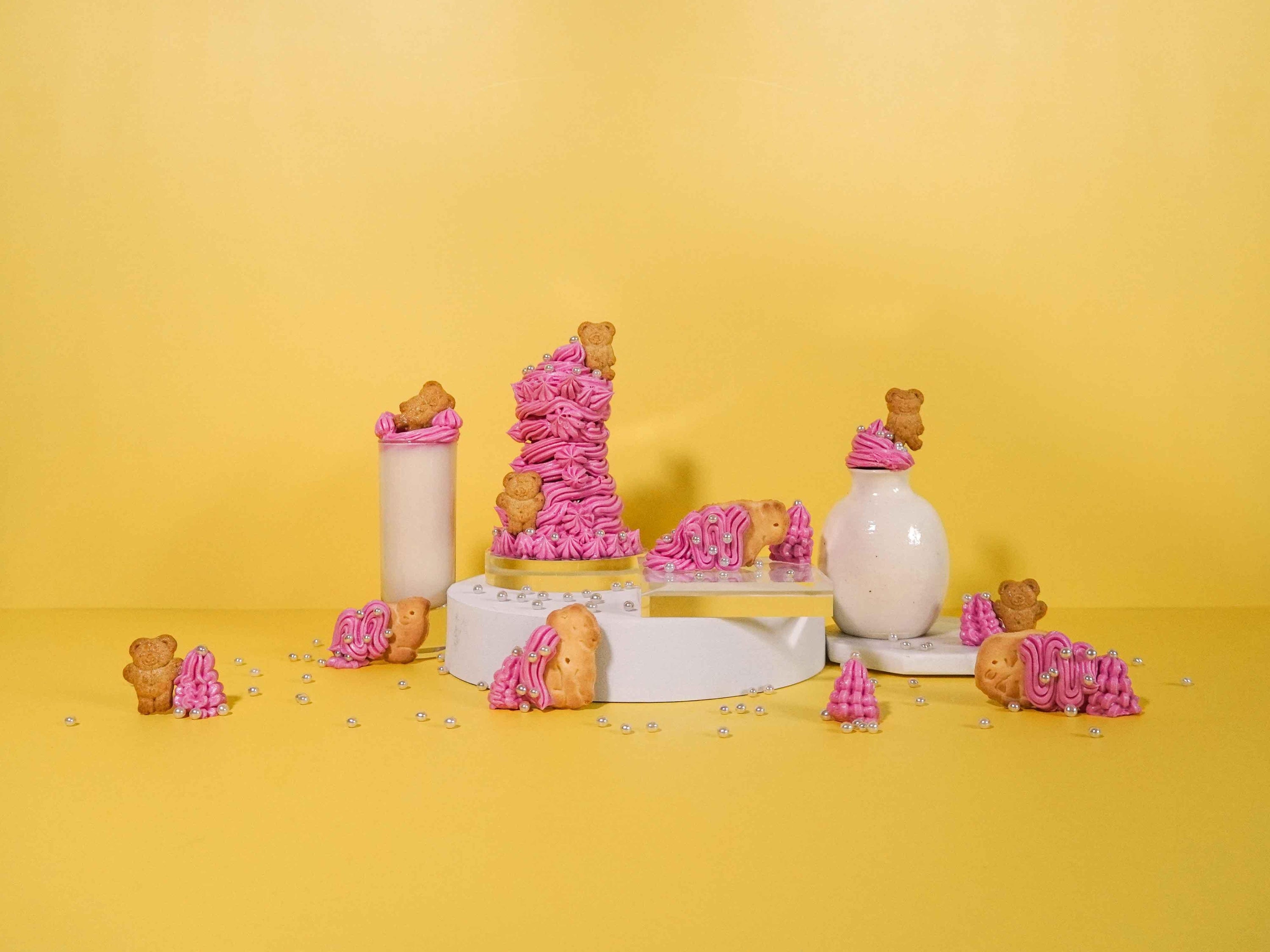 A whimsical arrangement featuring pink frosted animal cookies and golden teddy bear-shaped cookies, adorned with pearl-like sugar decorations. The scene includes a tall glass of milk and a simple white ceramic vase, all placed on a sunny yellow background. Playful stacks and scattered cookies create a cheerful, nostalgic atmosphere reminiscent of childhood treats.