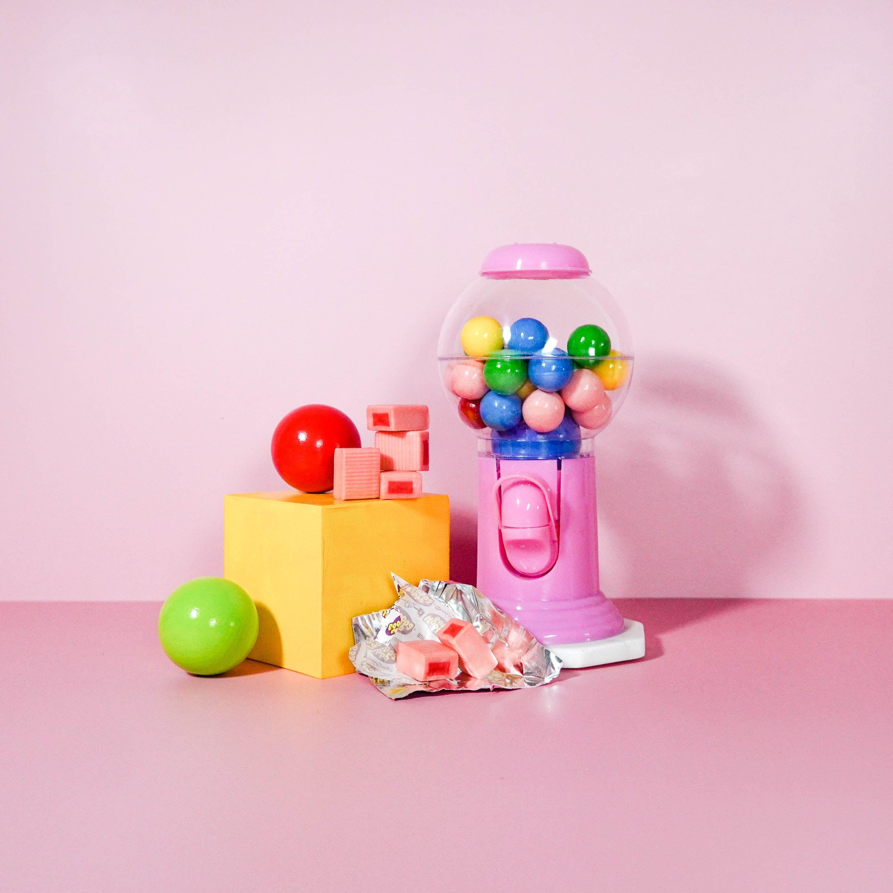 A colorful still life setup with a pink gumball machine, surrounded by various playful items like a yellow block, a green apple, and some wrapped candies.
